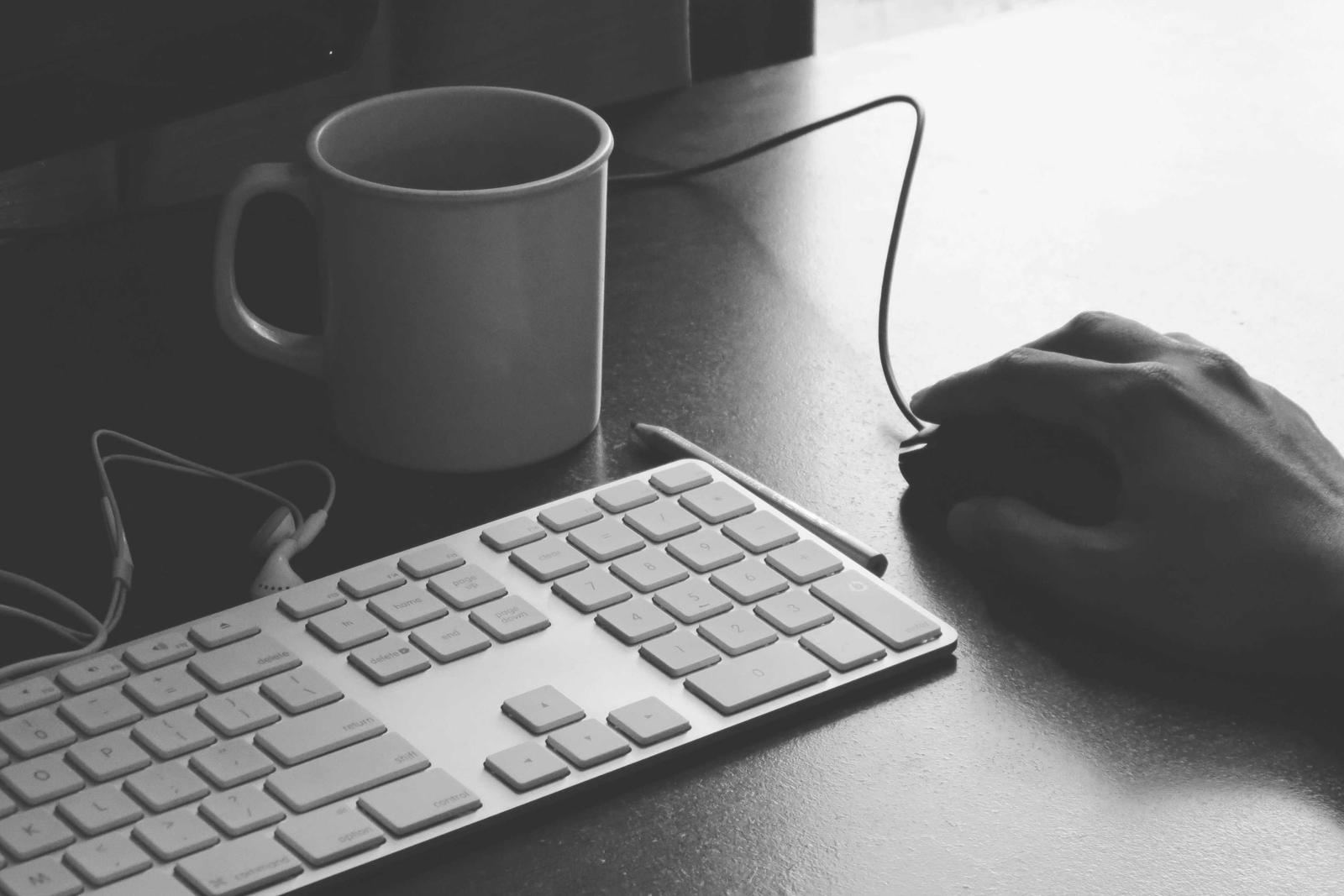 A hand on a mouse beside a keyboard and coffee mug